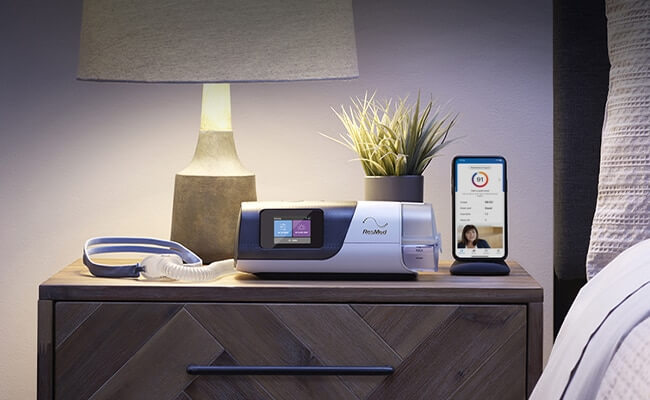 En Air11 sömnapné-apparat , mask och en mobiltelefon med myAir-appen på ett nattduksbord.