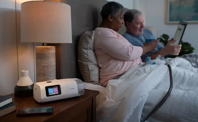 Ett par som ligger i sängen och tittar på en surfplatta. Kvinnan har en mask till en AirCurve 11 ASV PaceWave-apparat i knät. Masken är ansluten till apparaten som står på hennes nattduksbord.