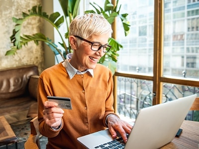 En äldre kvinna med kort hår och glasögon som onlineshoppar på sin laptop med sitt bankkort i ena handen